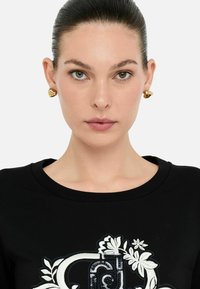Zwarte sweatshirt met ronde halslijn, voorzien van witte bloemembroideries en pailletten, gecombineerd met gouden hartvormige oorbellen. Gladde textuur.