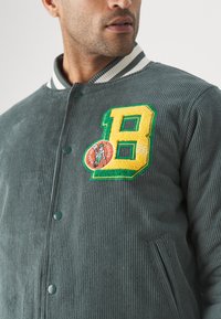 Giacca in corda verde con una texture a coste, dotata di un patch giallo con la lettera "B" e il logo dei Boston Celtics, bottoni a pressione e tasche laterali.