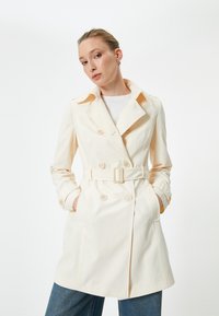 Trench coat beige con bottoni doppi, vita con cintura e maniche lunghe. Tessuto liscio con colletto classico e tasche laterali.