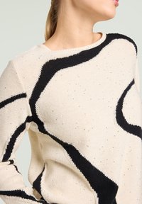 Maglione crema con motivo astratto nero e sottili scintillii argentati. Presenta un collo rotondo, maniche lunghe e tessuto a maglia strutturato.