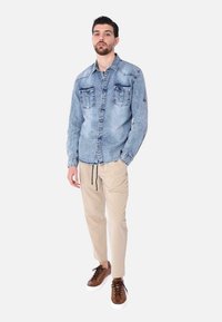 Camicia di jeans azzurro chiaro con due tasche sul petto, chiusura a bottoni e maniche lunghe. Abbinata a pantaloni beige e sneakers marroni.