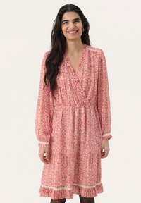 Mujer sonriendo, vestida con un vestido de manga larga y hasta la rodilla con un patrón floral rojo, con un dobladillo con volantes y encaje en los puños y en la parte inferior de la falda.