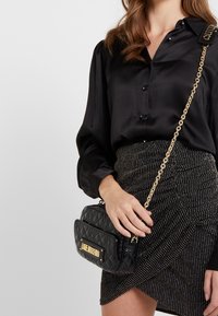 Svart vadderad handväska med en guldfärgad logoplåt och kedjesnor, som har en rektangulär form och texturerad yta. Bärs tillsammans med en svart blus och randig kjol.