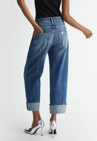 Světle modré rifle s vysokým pasem a ohrnutými lemy, s kapsami na zadní straně a kontrastním prošitím, doplněné stříbrnými botami na vysokém podpatku.