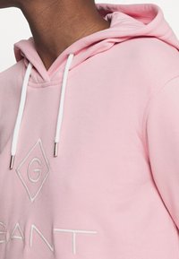 Sudadera con capucha rosa y cordones blancos, con un logo y letras bordadas en la parte delantera en un color contrastante.