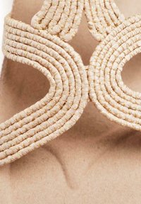 Sandal tejido beige con tiras curvadas de diseño trenzado, apoyado sobre una suela suave de tono neutro. Textura suave, con ornamentación mínima.