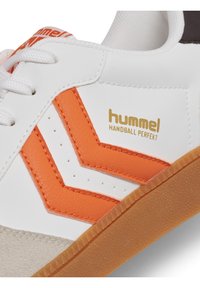Biały sneaker z pomarańczowym wzorem w kształcie chevron, wykonany ze skóry i zamszu, z fakturowaną gumową podeszwą oraz złotym logo "hummel".