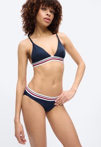 Femeie cu păr creț purtând un bikini bleu închis cu benzi elastice dungi albe, roșii și negre, stând cu o mână pe șold.