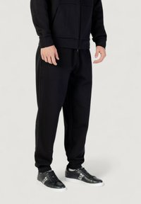 Schwarze Zip-Jacke mit gerippten Bündchen, kombiniert mit lässigen schwarzen Sweatpants. Die Hose hat elastische Knöchel-Bündchen. Schwarze Sneaker runden den Look ab.