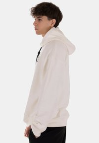 Felpa bianca con cappuccio e cordino, vestibilità rilassata e polsini a costine. Design minimalista con una piccola grafica sul davanti. Texture morbida.