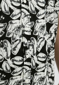 Kurzärmliges Hemd mit Knöpfen, das aus einem schwarzen Stoff mit weißem Blumenmuster besteht, ein strukturiertes Design aufweist und weiße Knöpfe entlang der Vorderseite hat.