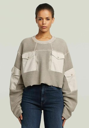 Giovane donna che indossa un maglione beige oversize con grandi tasche frontali e sulle maniche e jeans blu scuro a vita alta, in piedi davanti a uno sfondo semplice.