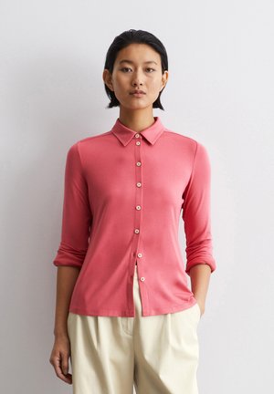 Chemise longue à manches roses avec col, fermeture à boutons sur le devant et manches retroussées. Fabriquée en matériau doux, elle présente une coupe décontractée et un subtil éclat.