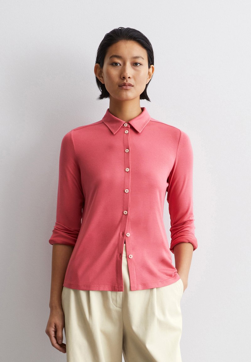 Camisa rosa de manga larga con cuello, parte delantera con botones y mangas enrolladas. Fabricada con un material suave, presenta un corte holgado y un brillo sutil.