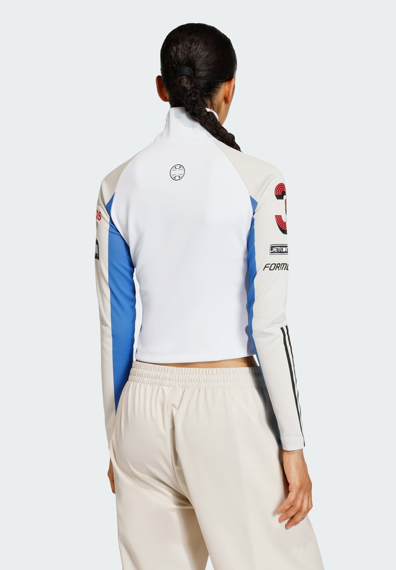 Kvindelig med fletninger iført en hvid, blå og beige langærmet bluse med logoer og beige bukser, vist fra ryggen.