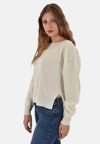 Maglione beige chiaro lavorato a maglia con texture a coste, scollo rotondo, maniche larghe e spacchi laterali. Abbinato a jeans aderenti blu scuro.