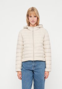 Beige pufferjack met een horizontaal gestikte patroon, capuchon en frontale rits. Zachte stof en cropped ontwerp in combinatie met blauwe jeans.