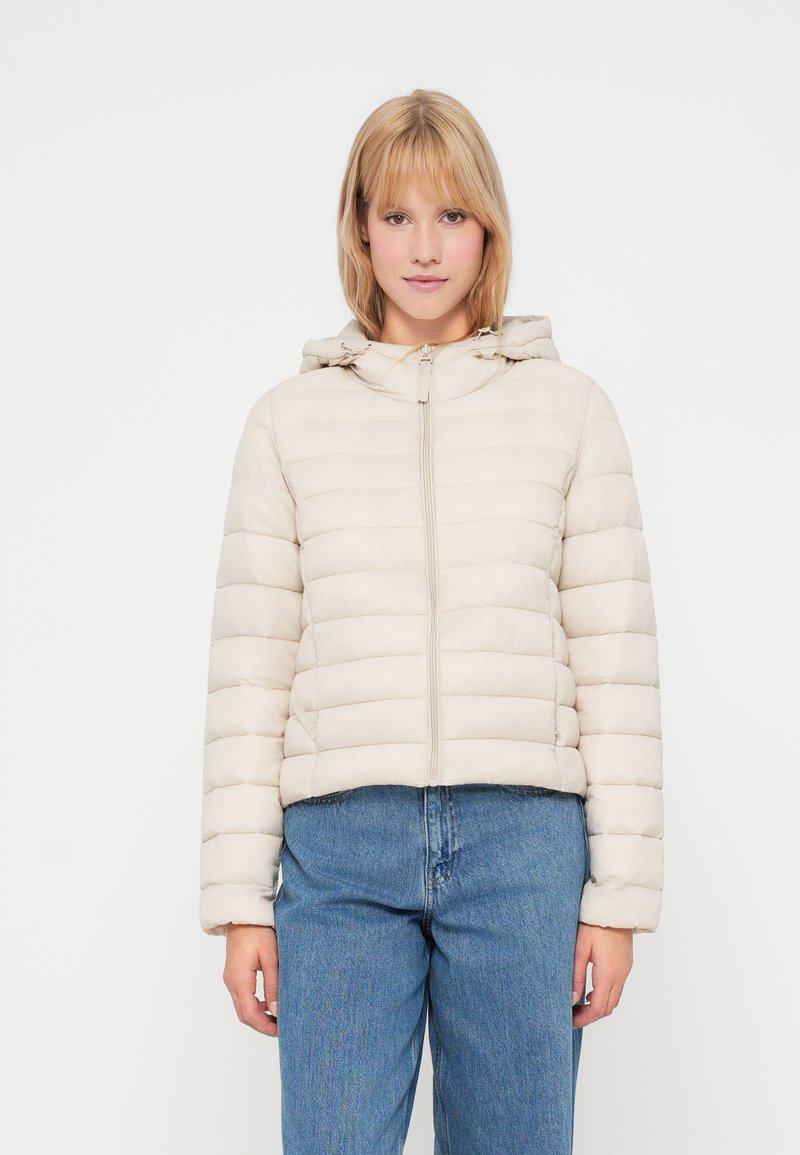 Beige pufferjack met een horizontaal gestikte patroon, capuchon en frontale rits. Zachte stof en cropped ontwerp in combinatie met blauwe jeans.