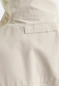 Lichtbeige jas stof met een soepele textuur en een GANT logo label op de achterkant gestikt. Het label heeft kruissteekaccenten.
