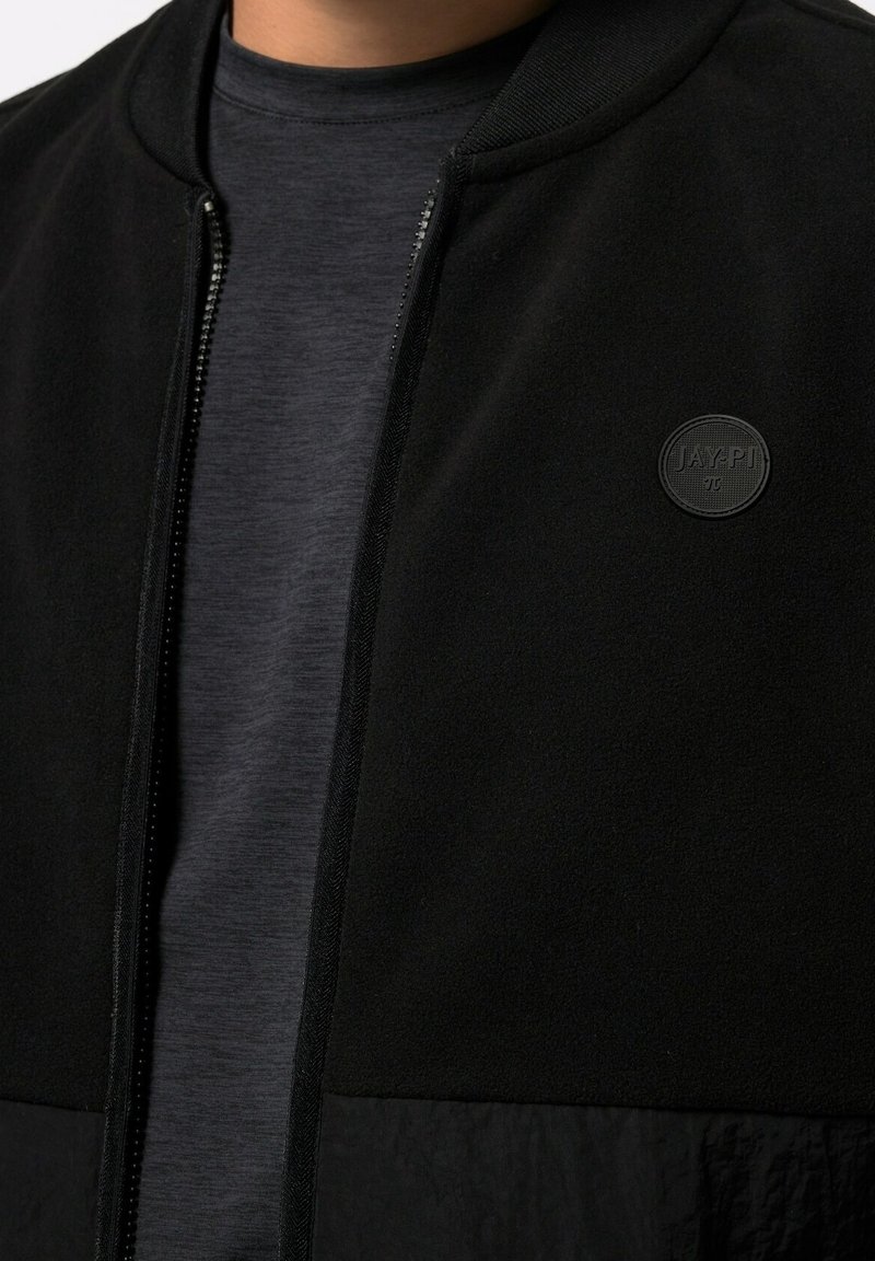 Gros plan d'une personne portant une chemise gris foncé sous une veste noire zippée avec un patch rond au logo « JAY.PI » sur la poitrine.