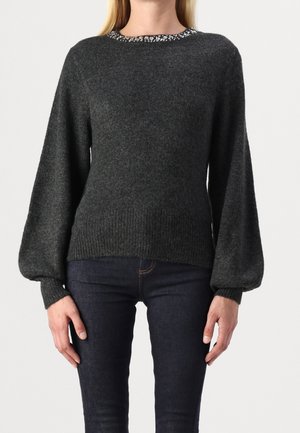 Femme portant un pull en maille gris foncé avec des manches bouffantes et un col orné, associée à un jean bleu foncé, debout, mains détendues.