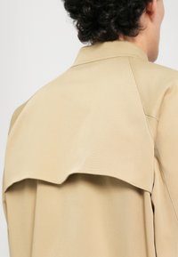 Moški s temnimi kodrastimi lasmi, oblečen v bež trench plašč, prikazan s hrbta, s poudarkom na detajlih zgornjega dela hrbta in ovratniku.