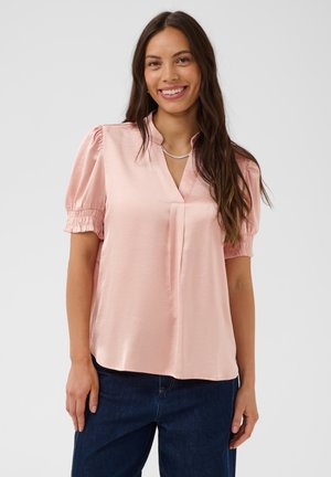 Femme souriante aux longs cheveux foncés portant un chemisier en satin rose clair avec des manches courtes bouffantes et un jean bleu foncé sur un fond uni.