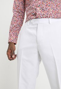 Pantalones blancos a medida con una textura suave y pliegues marcados, combinados con una camisa de estampado floral multicolor con botones y puños.