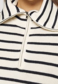 Gros plan sur un pull blanc en maille avec des rayures horizontales noires et une fermeture éclair argentée partiellement zippée au col montant.