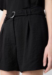 Shorts plissés noirs avec une surface texturée et une ceinture en tissu dotée d'une boucle en D en métal argenté, présentés de la taille vers le bas avec une main visible.