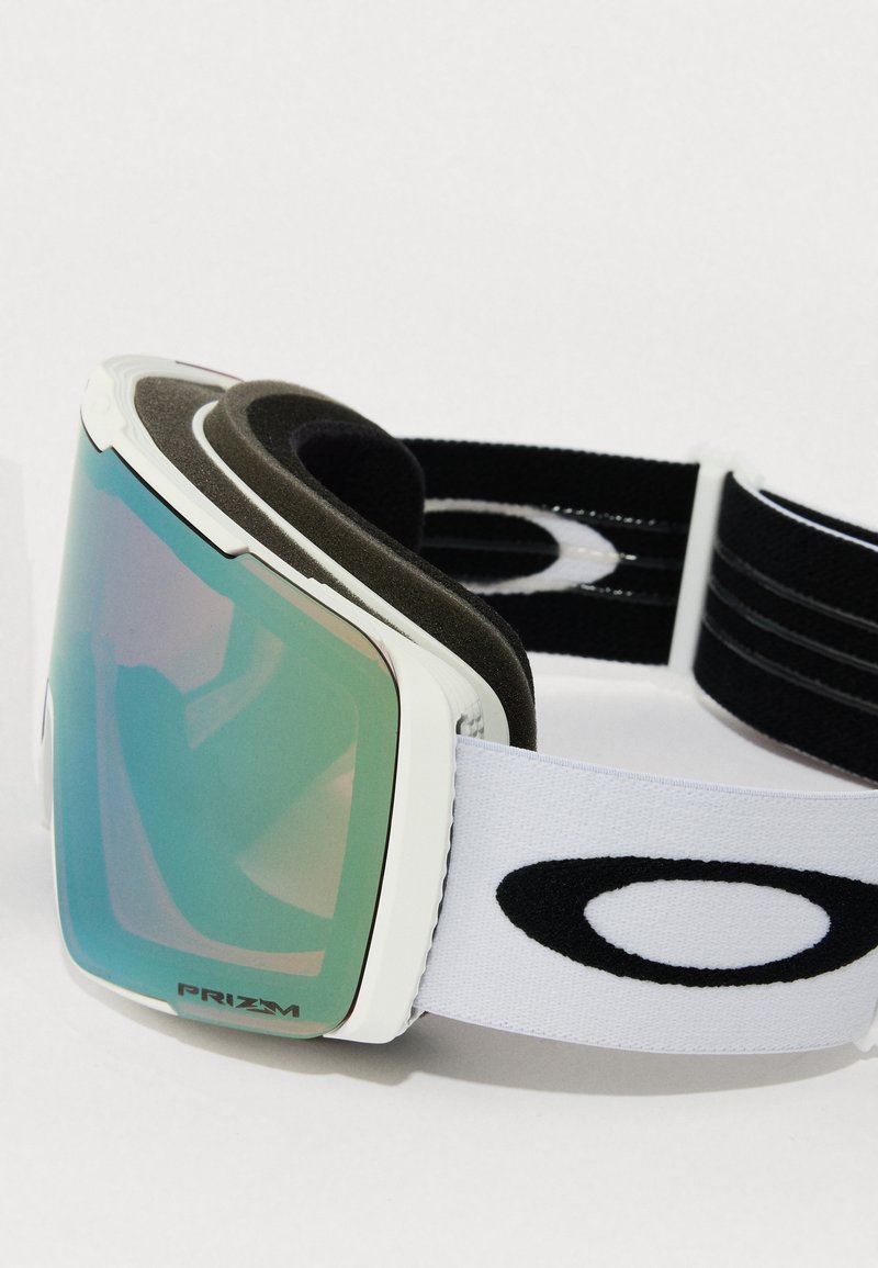 Skibril met een wit frame, groen getinte lens en zachte zwarte schuimrubberen padding. Heeft een witte elastische band met een zwart logo.