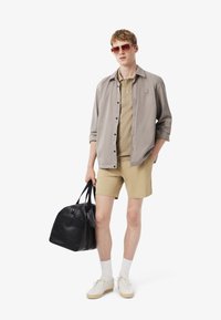 Ljusgrå knapp-up skjorta över en beige polo, i kombination med beige shorts. Svart duffelväska och vita sneakers med beige sålor. Bruna solglasögon.