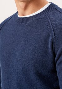 Gros plan sur une personne portant un pull en tricot bleu marine avec un t-shirt blanc à col rond en dessous, montrant les détails de l'épaule et du cou.
