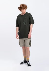 Donkergroen katoenen T-shirt met een eenvoudig lijntekeningontwerp, gecombineerd met beige cargoshorts met zijzakken en zwarte sneakers.