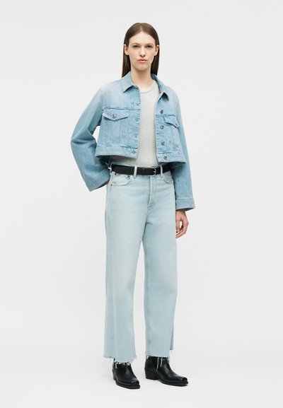 Γυναίκα με κοντό ανοιχτό μπλε τζιν σακάκι, λευκή μπλούζα, τζιν παντελόνι ανοιχτό μπλε ψηλόμεσο με φθαρμένα τελειώματα, μαύρη ζώνη και μαύρα μποτάκια μέχρι τον αστράγαλο.