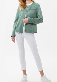 Grön mockajacka med rund halsringning, knappframsida och långärmade, kombinerad med vita åtsittande byxor och vita sneakers.