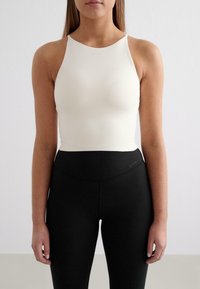 Top sportivo bianco a collo alto in tessuto elasticizzato, con spalline sottili, abbinato a leggings neri a vita alta con un branding discreto.