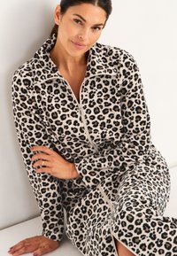 Leopardmönstrad dragkedjesydd fleeceplagg med långa ärmar, mjuk textur och kontrasterande beige bas. Modellen sitter mot en vit bakgrund.