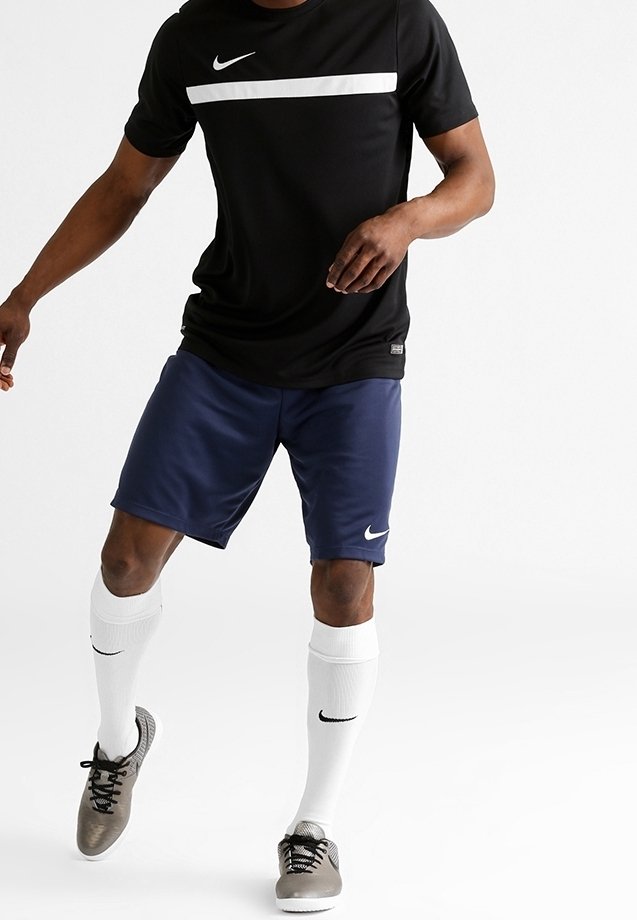 Homme portant un t-shirt de sport Nike noir, un short Nike bleu marine, des chaussettes blanches Nike et des crampons gris, debout sur fond blanc.