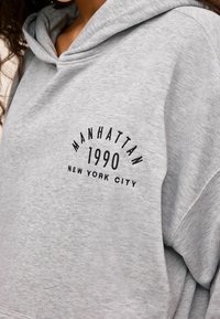 Sudadera gris con capucha, con texto negro: "MANHATTAN 1990 NUEVA YORK" en un diseño arqueado en la parte frontal. Textura de material suave.