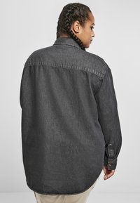 Svart denimskjorte med løs passform, knappeermer og spiss krage. Teksturen virker jevn med en lett glans. Vist bakfra.