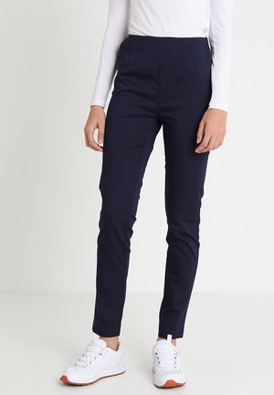 Person iført tætsiddende marineblå bukser, hvid top med lange ærmer og hvide sneakers med orange såler, stående mod en ensfarvet baggrund.