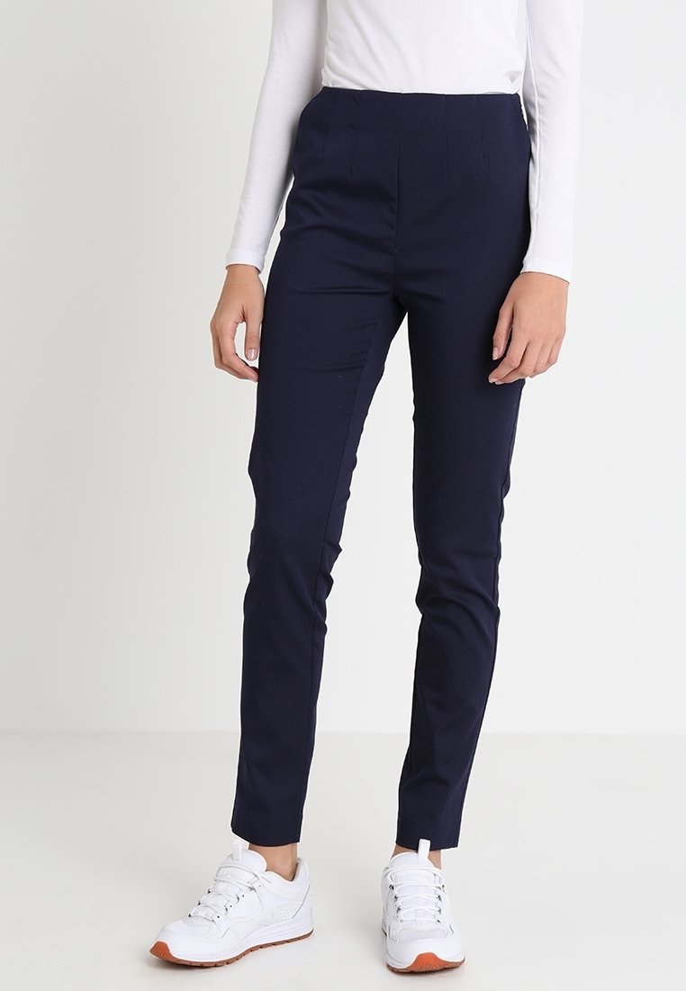Personne portant un pantalon bleu marine slim, un haut blanc à manches longues et des baskets blanches avec des semelles orange, debout devant un fond uni.