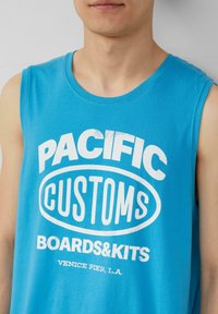 Niebieska koszulka bez rękawów wykonana z bawełny, z wyraźnymi białymi grafikami tekstowymi: "PACIFIC CUSTOMS BOARDS&KITS" oraz "VENICE PIER, L.A."