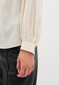 Blusa di un leggero color crema con tessuto testurizzato, caratterizzata da maniche lunghe a palloncino e dettagli di polsino/bottoni ai polsi. Abbinata a pantaloni in pelle nera.