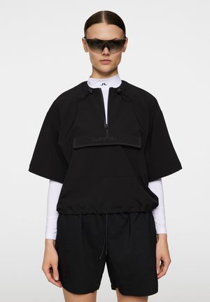 Zwarte pullover met korte mouwen, hoge kraag, half-rits aan de voorkant en een voorzak. Gecombineerd met aansluitende witte lange mouwen en zwarte shorts.