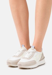 Białe i beżowe sneakersy z teksturowaną zamszową i gładką syntetyczną cholewką. Posiadają białe sznurówki, szarą podeszwę oraz jasny żółty akcent w designie.