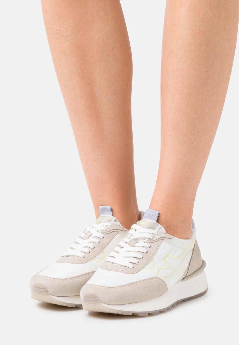 Białe i beżowe sneakersy z teksturowaną zamszową i gładką syntetyczną cholewką. Posiadają białe sznurówki, szarą podeszwę oraz jasny żółty akcent w designie.