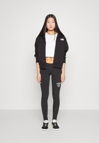 Čierna zipová bunda, biely crop top, čierne legíny s bielym logom Adidas a čierne tenisky so žltými akcentmi. Jednoduché pozadie.