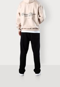 Beżowa bluza z kapturem z czarnym napisem na plecach, zestawiona z czarnymi spodnie dresowymi, subtelna faktura i białe trampki. Minimalistyczny design.